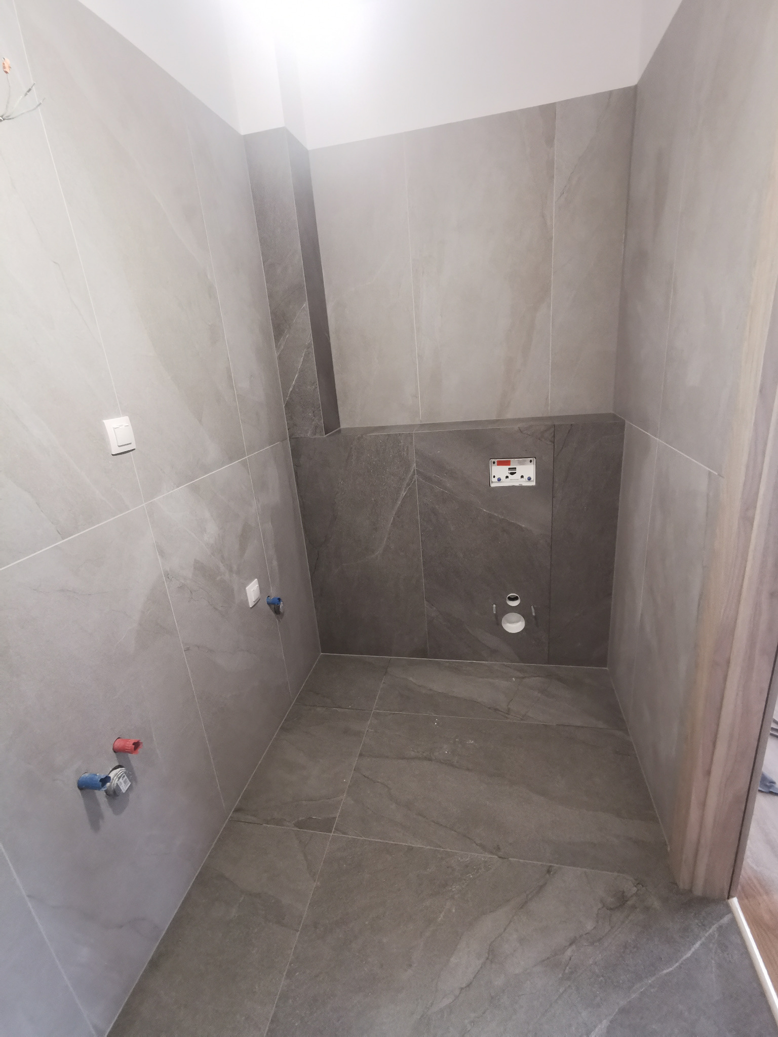 Wykończona łazienka w odcieniach szarości, z widocznymi przyłączami wodnymi i instalacją podtynkową WC, płytki imitujące kamień na ścianach i podłodze, minimalistyczny design.