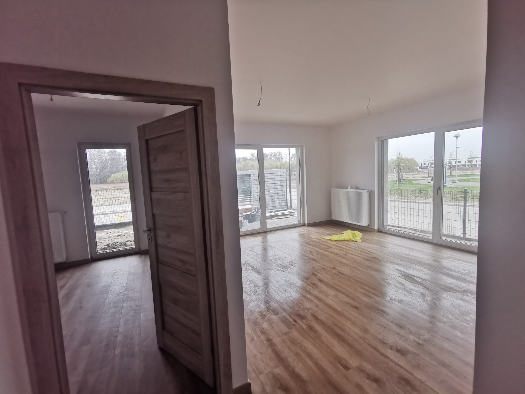 Wnętrze nowego mieszkania z drewnianą podłogą, widok przez otwarte drzwi z szarą ościeżnicą na kolejny pokój, w tle okna z widokiem na plac budowy i park.