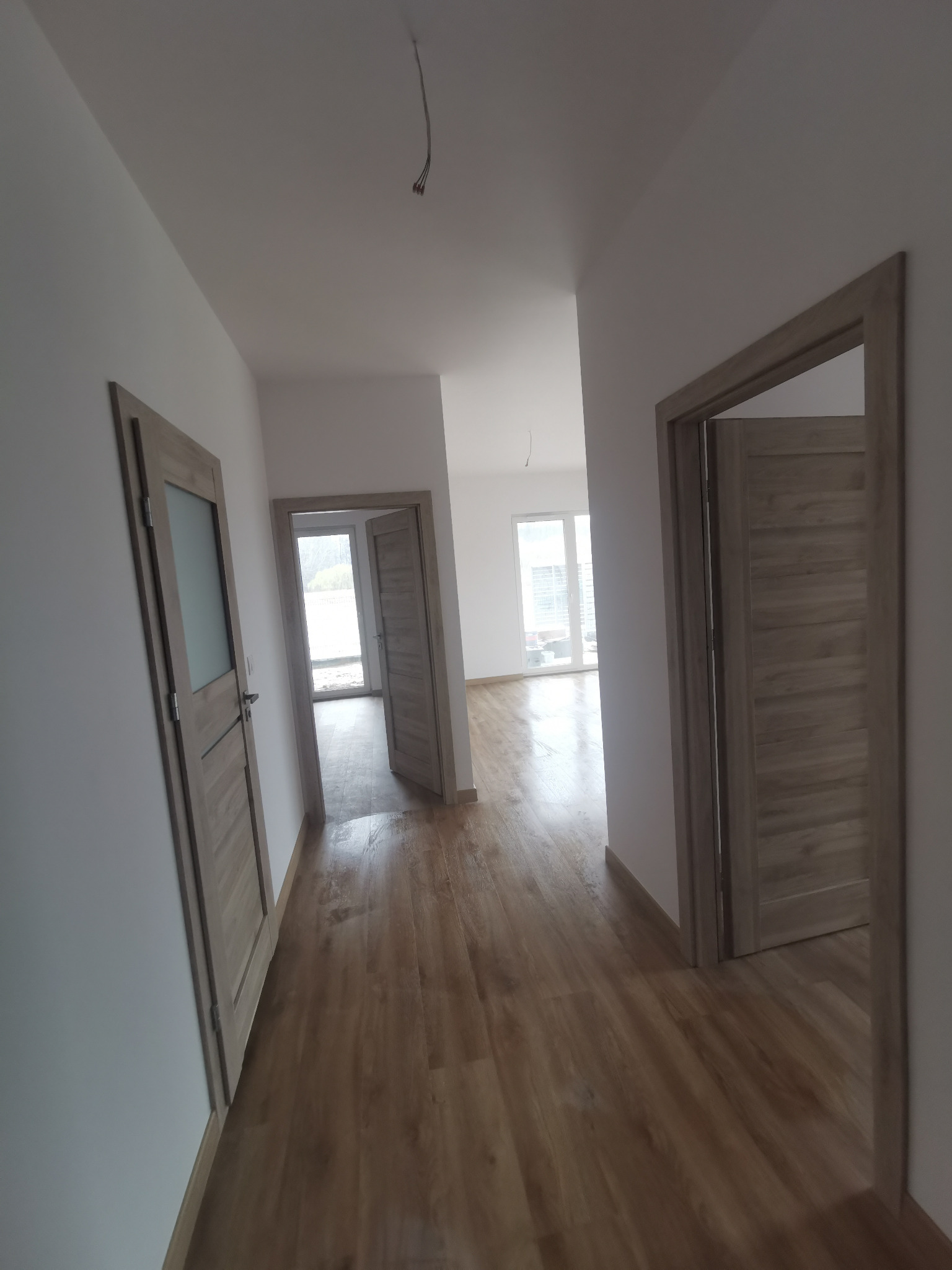 Wnętrze wykończonego mieszkania: drewnopodobna podłoga, białe ściany, zamontowane drzwi wewnętrzne w odcieniach szarości z widocznym usłojeniem, oraz częściowo przeszklone drzwi po lewej stronie.