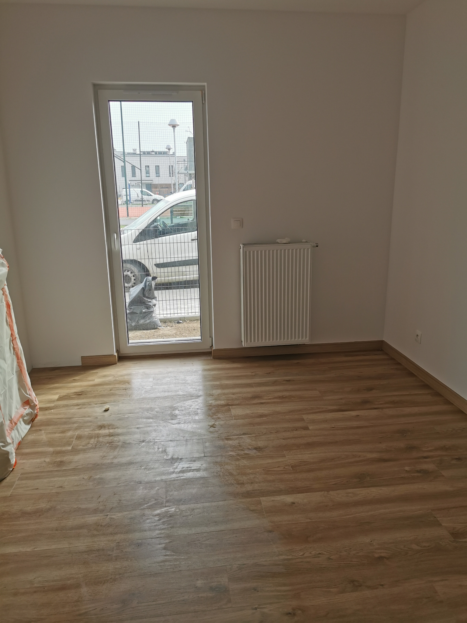 Pusty pokój po remoncie z panelami podłogowymi w odcieniach brązu, grzejnikiem na ścianie i widokiem na ulicę przez okno, z workiem gruzu za oknem.