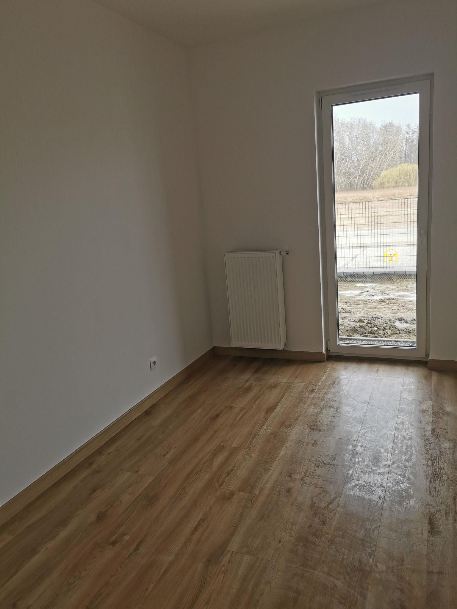 Pusty pokój z panelami podłogowymi, grzejnikiem na ścianie i widokiem na plac budowy za oknem.