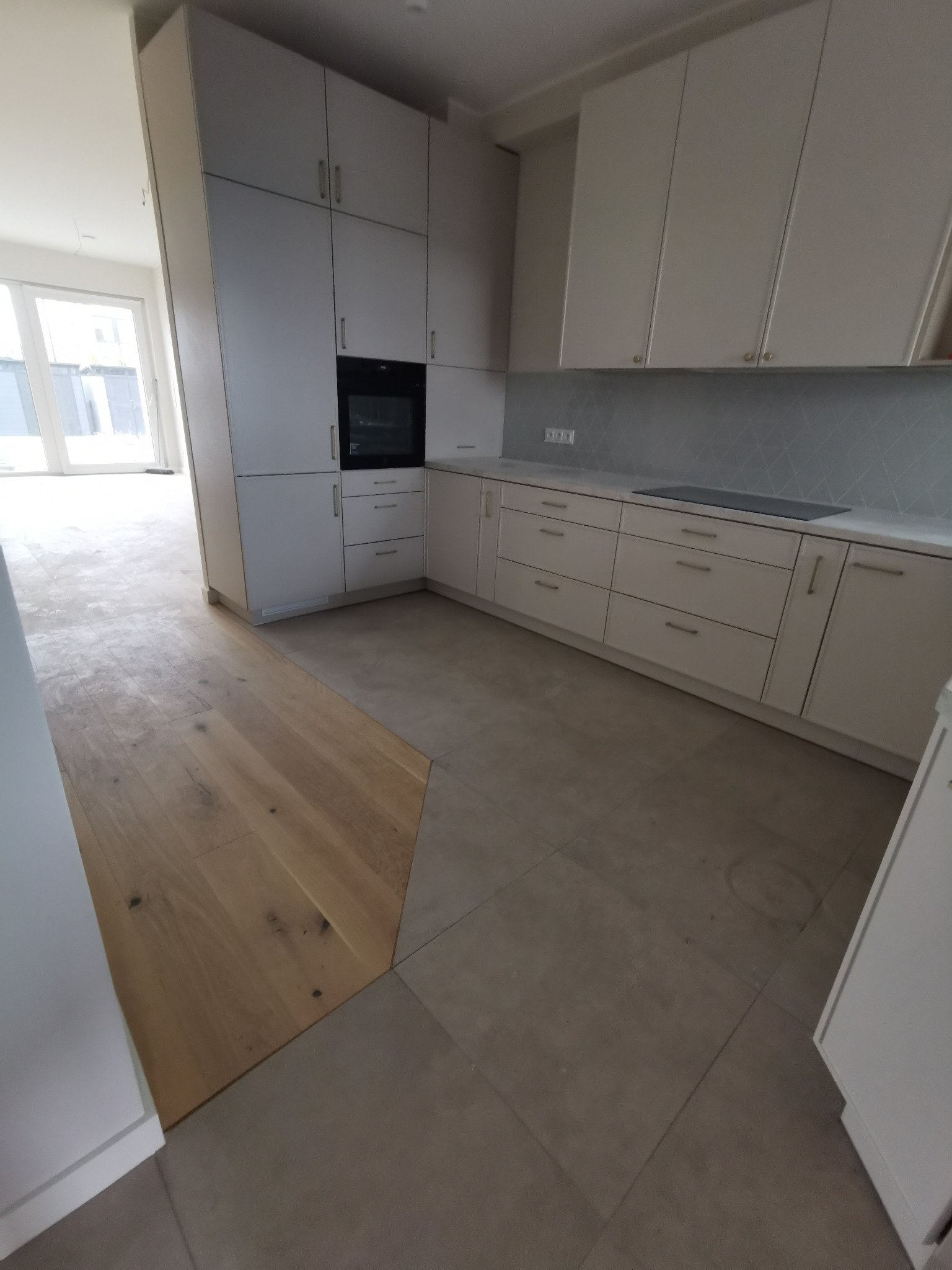 Jasna kuchnia z zabudową meblową, drewnianą podłogą przechodzącą w szarą płytkę, widoczny fragment salonu z oknami balkonowymi.