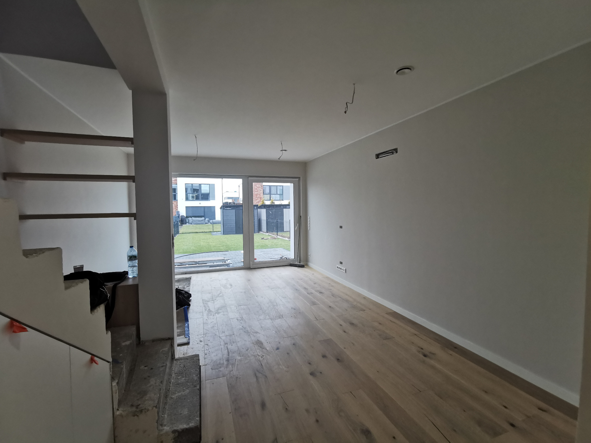 Jasne, puste pomieszczenie z nową, drewnianą podłogą i surowymi schodami w trakcie wykończenia, widok na ogród przez przeszklone drzwi przesuwne.