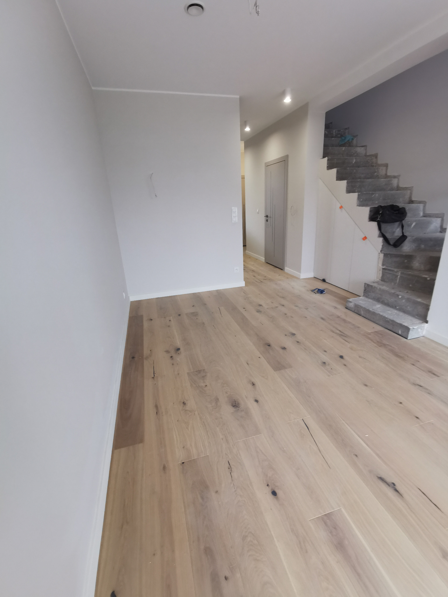 Jasna, drewniana podłoga z widocznymi sękami w nowym domu, z surowymi schodami betonowymi w tle i ścianami przygotowanymi do malowania.