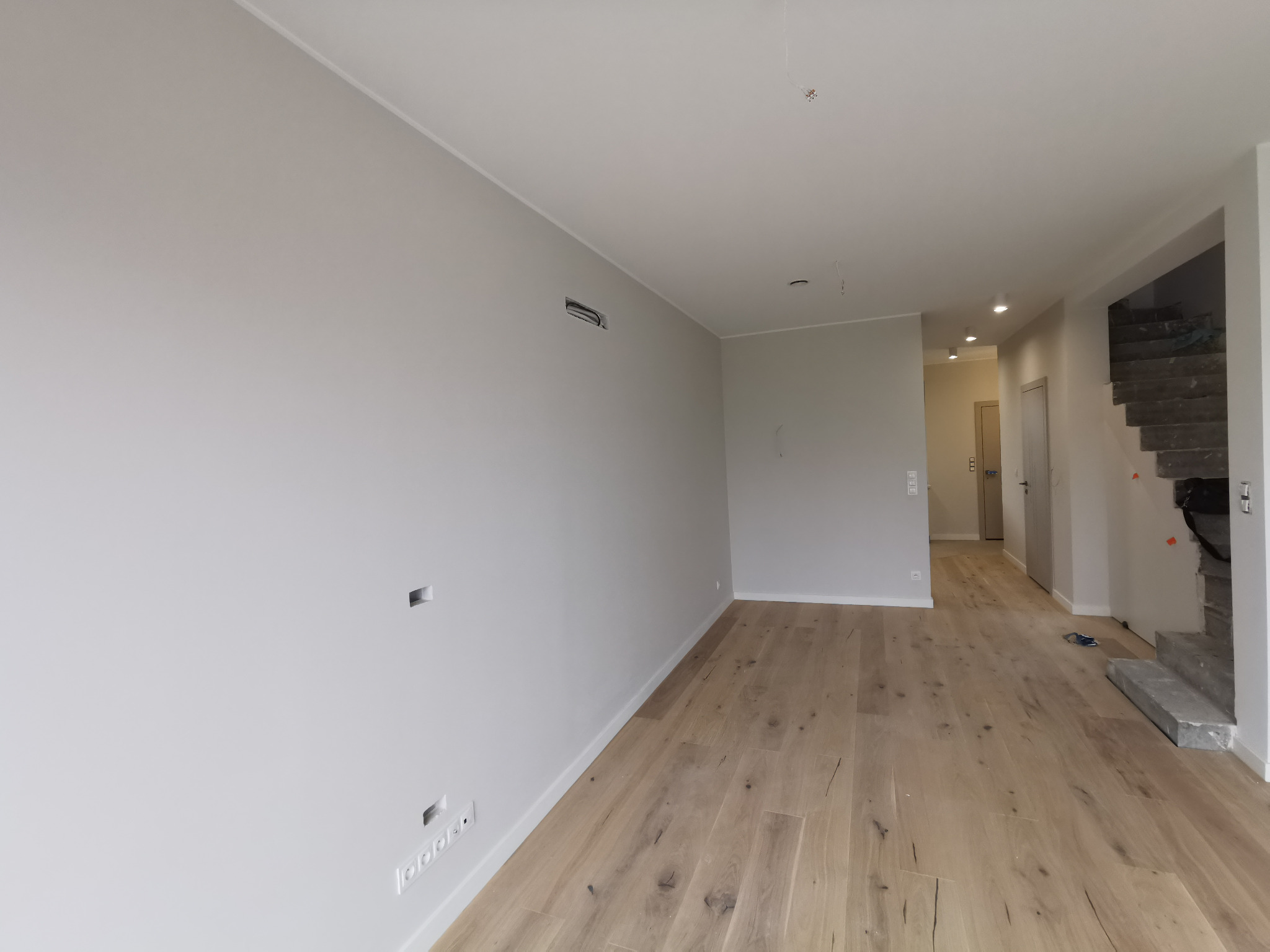 Wnętrze w Poznaniu po remoncie: świeżo pomalowane ściany w odcieniach szarości, jasna drewniana podłoga z widocznymi słojami, przygotowane otwory na gniazdka elektryczne i fragment schodów...