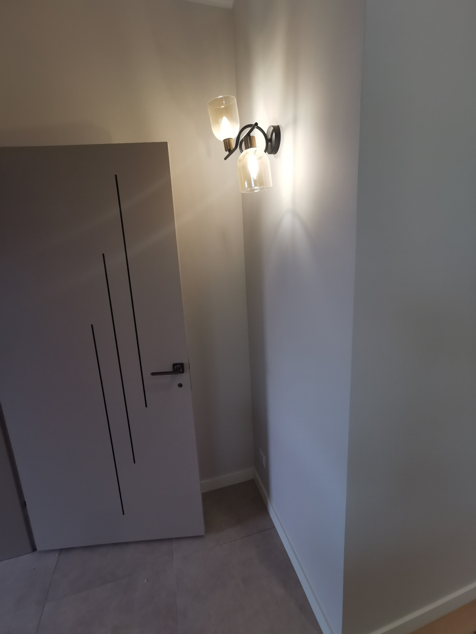 Szare drzwi z czarnymi, pionowymi zdobieniami i czarną klamką, oświetlone przez nowoczesny kinkiet z dwoma szklanymi kloszami w odcieniach beżu, na tle szarej ściany i podłogi z płytek.