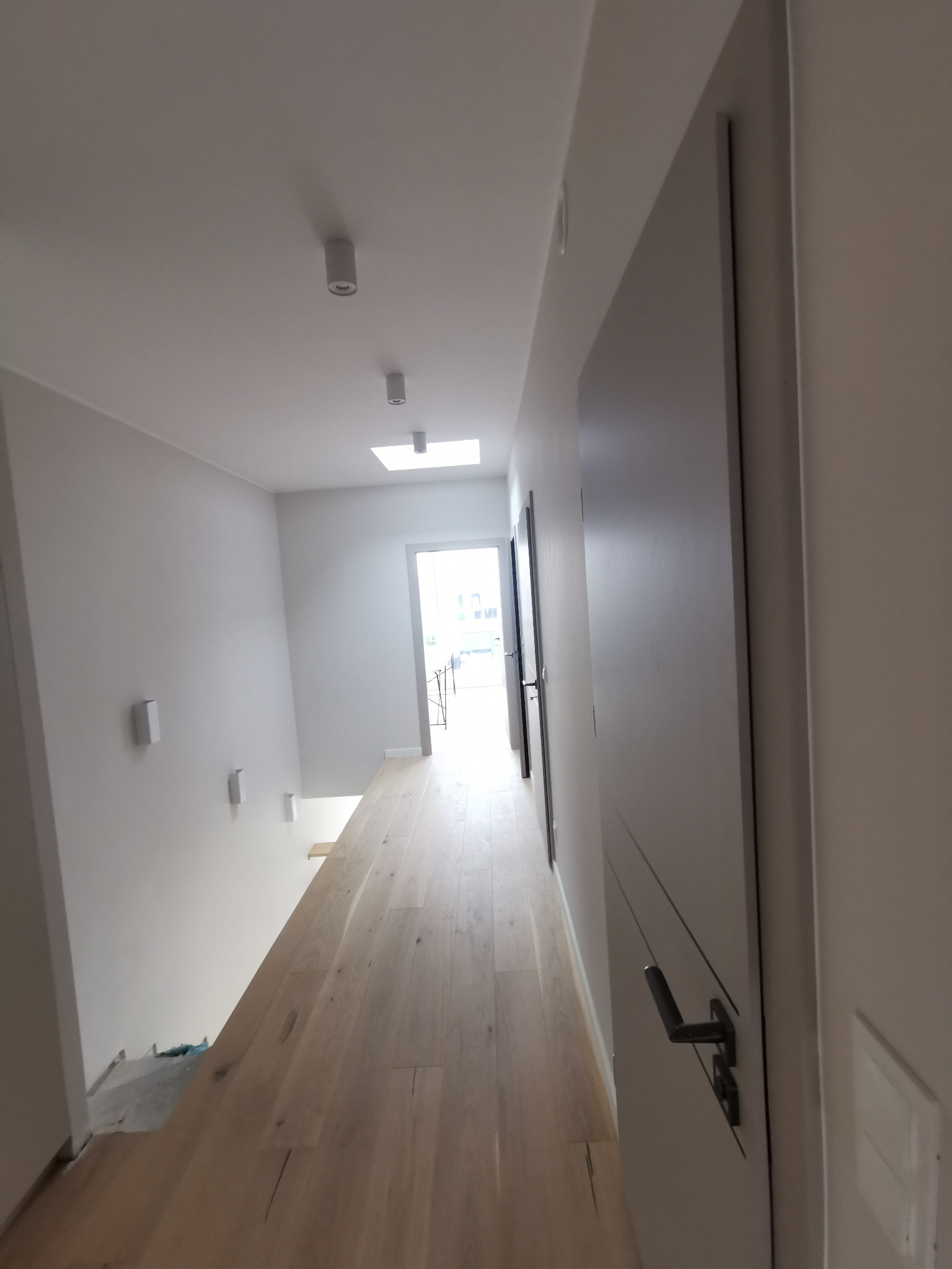 Nowoczesny korytarz z jasną drewnianą podłogą, minimalistycznymi lampami sufitowymi i białymi ścianami. Widoczne drzwi wewnętrzne z czarnymi klamkami i delikatnymi frezowaniami. W oddali widoczne...