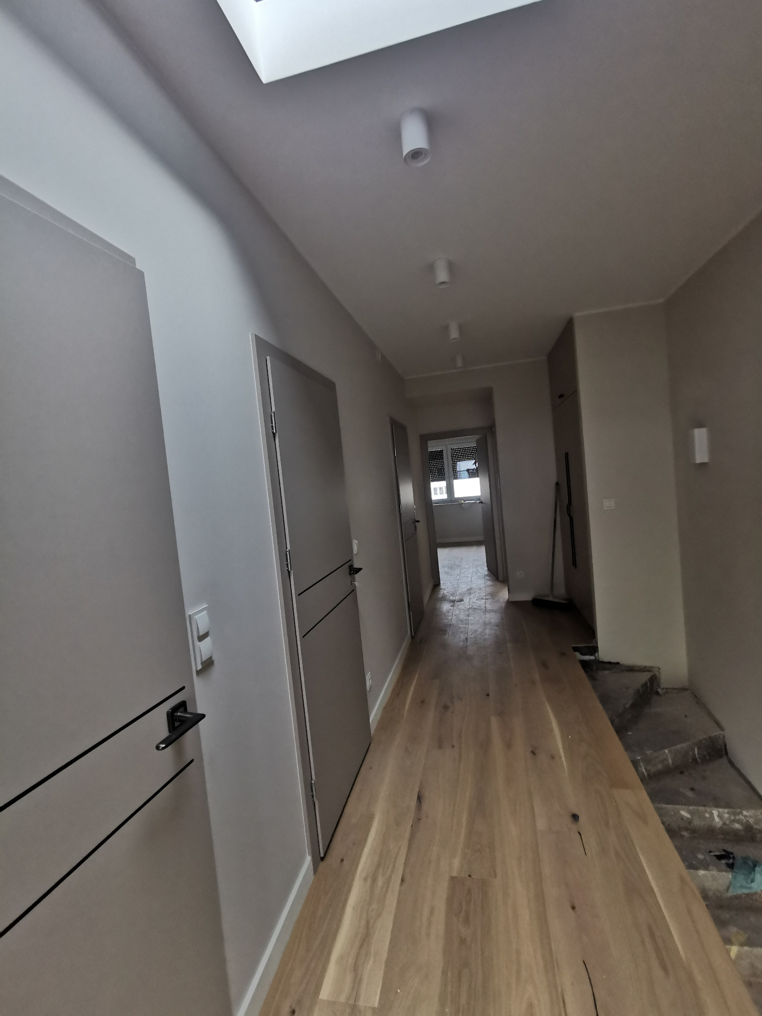 Długi korytarz po remoncie z nową, jasną drewnianą podłogą, szarymi drzwiami z czarnymi klamkami i białymi ścianami, widoczne schody w trakcie wykończenia.
