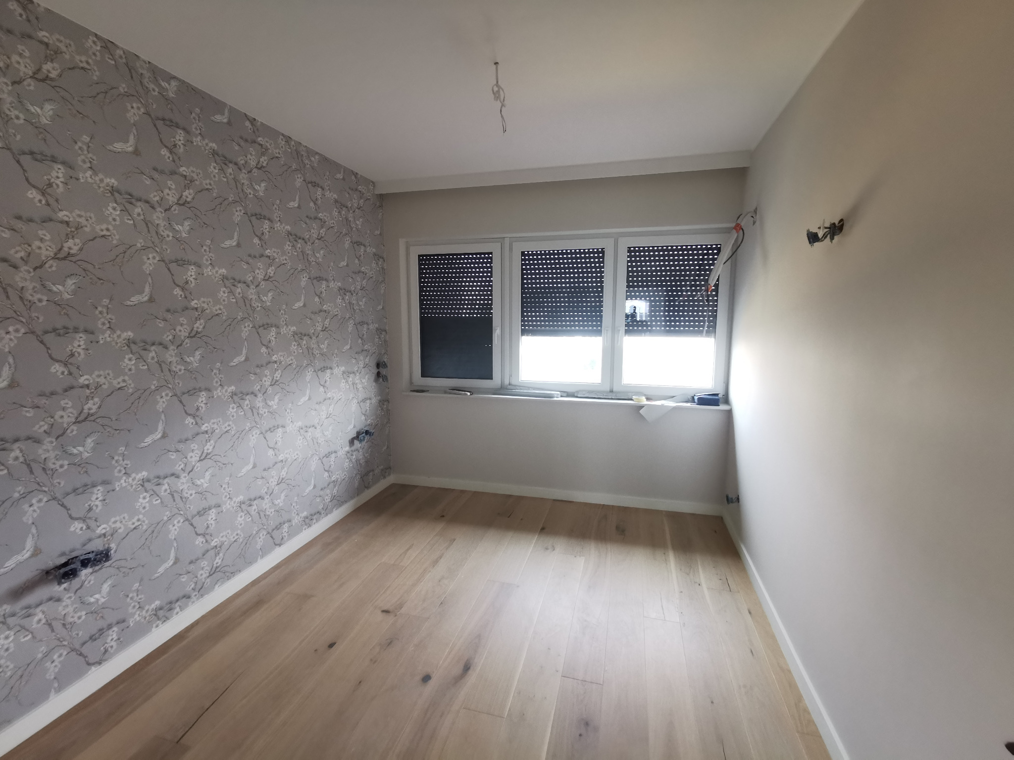 Pokój po remoncie z jasną drewnianą podłogą, ścianą w kwiatowy wzór i świeżo pomalowaną ścianą w kolorze kremowym, z widocznymi puszkami elektrycznymi i oknem z roletami.