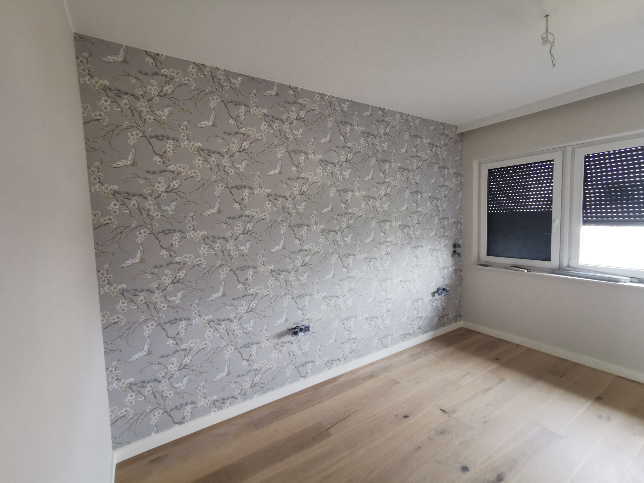 Pokój po remoncie z wzorzystą tapetą w odcieniach szarości z motywem kwitnących gałęzi i ptaków, drewnianą podłogą i białymi listwami przypodłogowymi, widoczne gniazdka elektryczne bez osprzętu.