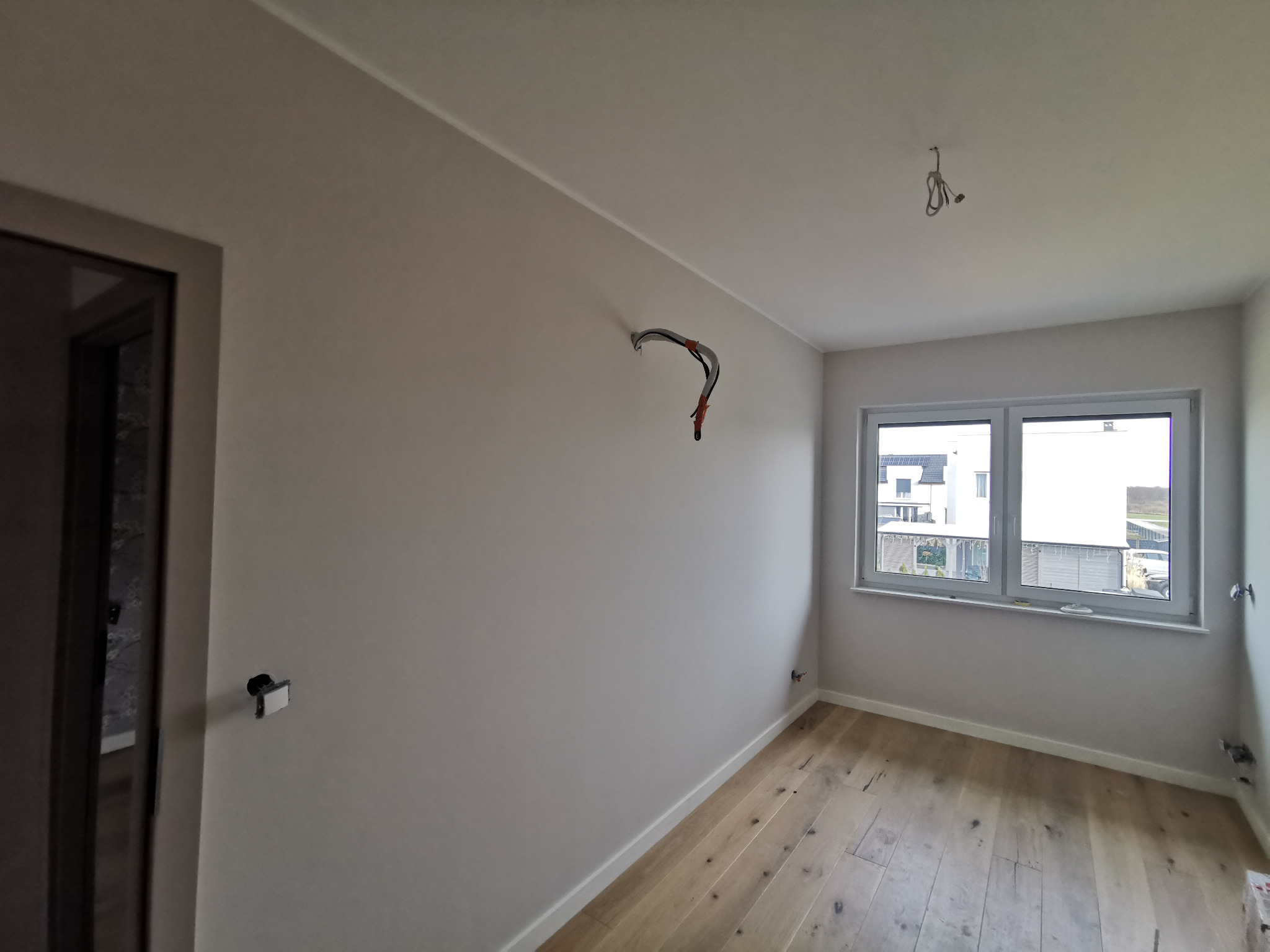 Pokój w trakcie remontu, z położonym parkietem, świeżo pomalowanymi ścianami na szaro, wystającymi przewodami elektrycznymi ze ścian i sufitu, oraz widocznym oknem z widokiem na domy.