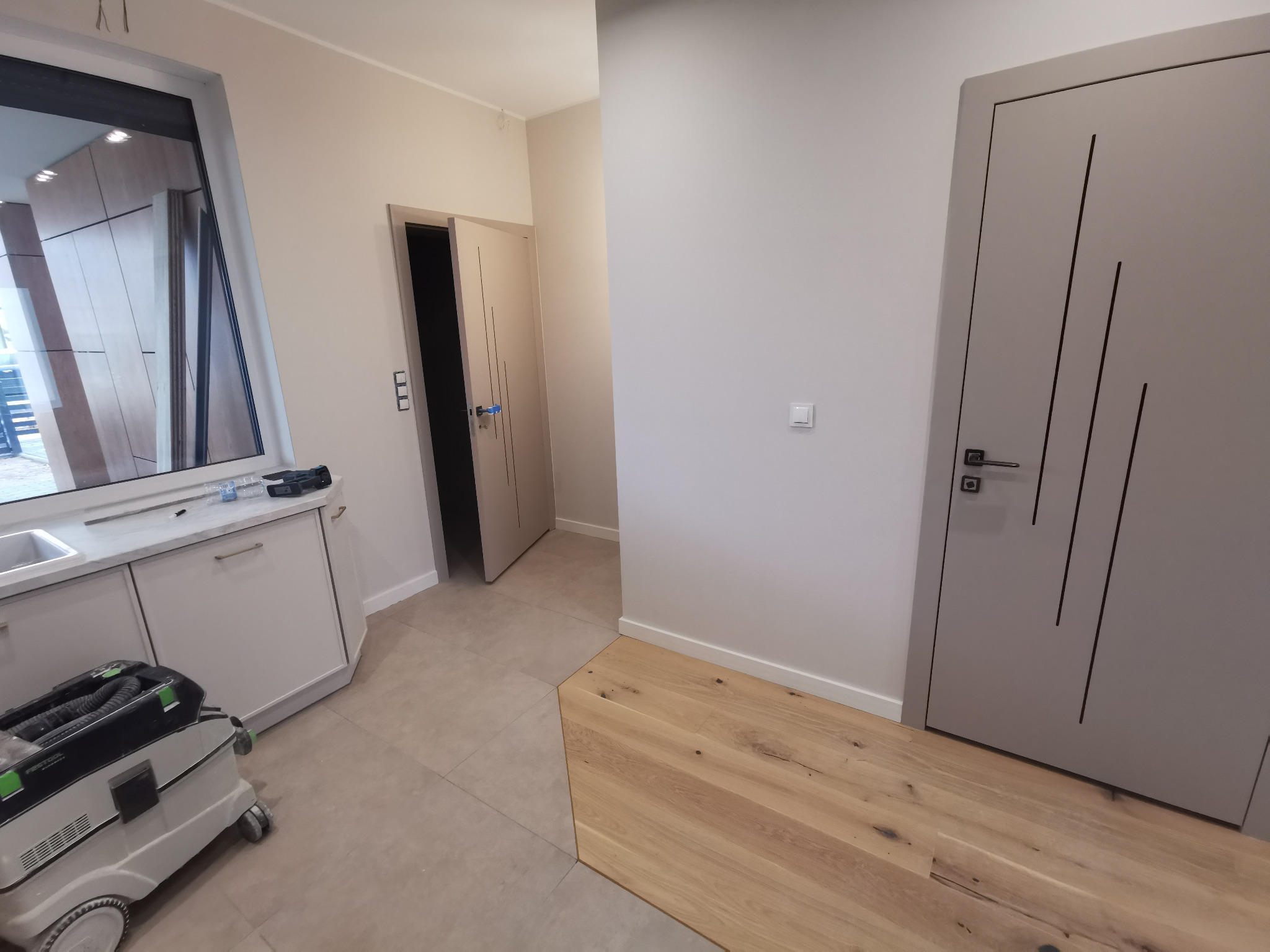 Jasne wnętrze po remoncie: widok na dwie szare drzwi z czarnymi wstawkami, fragment drewnianej podłogi przechodzącej w płytki, białe szafki pod oknem i odkurzacz przemysłowy.
