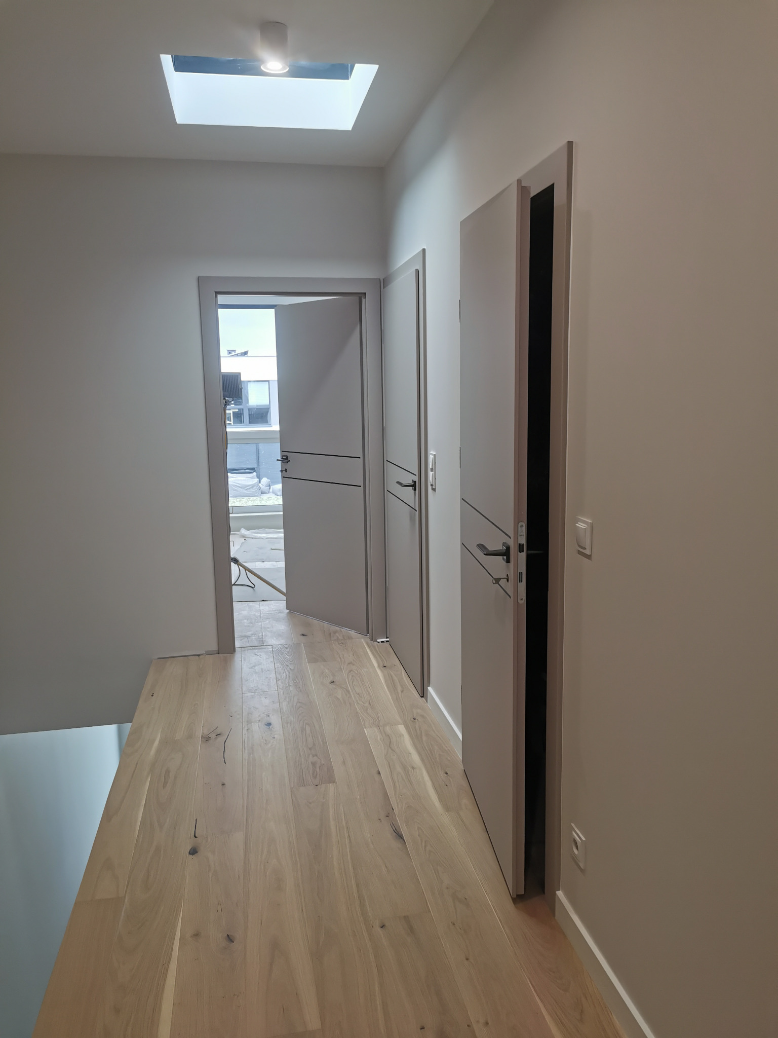 Korytarz w jasnych barwach z drewnianą podłogą, dwoje szarych drzwi z poziomymi zdobieniami, jedno drzwi otwarte, w suficie okno dachowe z oświetleniem.