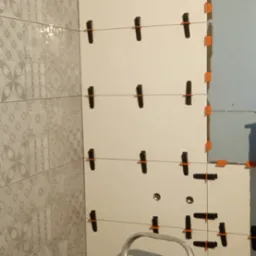 Układanie płytek ściennych w łazience, widoczne poziomice i krzyżyki dystansowe, częściowo ułożone płytki z wzorem, drabina aluminiowa w kadrze.