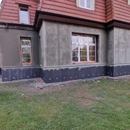 BUD-MAR - Dom w trakcie ocieplania: widoczne szare ściany po szpachlowaniu, czarne płyty izolacyjne przy fundamencie, okna zabezpieczone folią, pracownik na rusztowaniu przygotowuje ścianę pod malowanie.