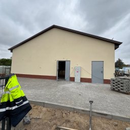 BUD-MAR - Budynek po malowaniu elewacji z widocznymi detalami: ceglana podmurówka, szara kostka brukowa, żółta kurtka i narzędzia sugerujące prace wykończeniowe na zewnątrz.