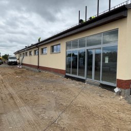 BUD-MAR - Budynek z beżową elewacją i dużymi oknami, z pracownikami na dachu, w trakcie prac wykończeniowych. Widoczny podjazd z piaskiem i ślady opon.