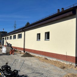 BUD-MAR - Prace wykończeniowe elewacji budynku: ocieplenie, ceglana podmurówka, żółta ściana. Na pierwszym planie ubijarka. Dwóch robotników przy pracy. Słoneczny dzień, błękitne niebo.