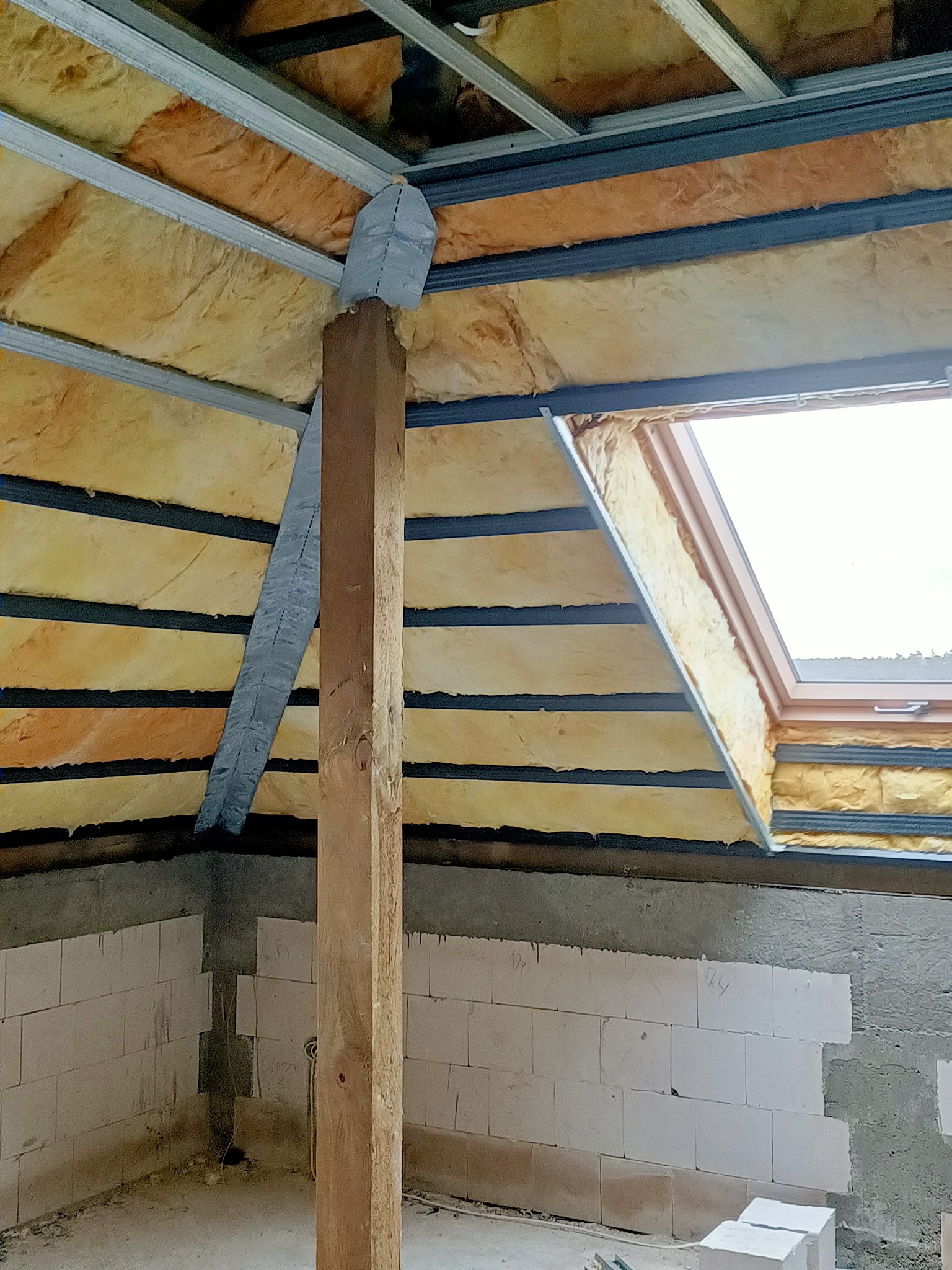 Izolacja poddasza wełną mineralną, widoczne metalowe profile, drewniany słup podtrzymujący konstrukcję, okno dachowe i fragment ściany z bloczków.
