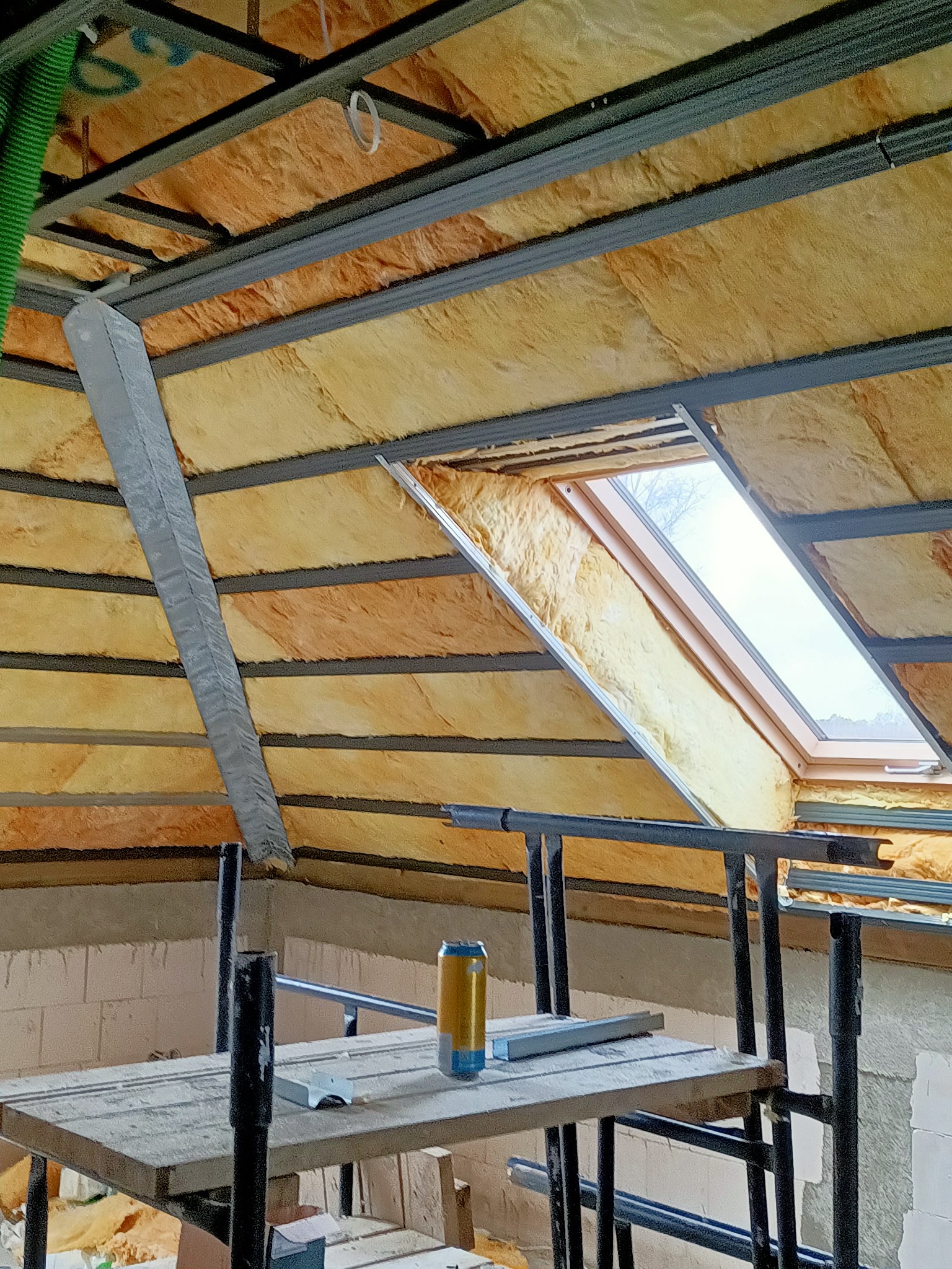 Montaż izolacji z wełny mineralnej na poddaszu z oknem dachowym, widoczne metalowe profile i rusztowanie pomocnicze oraz puszka napoju na prowizorycznym stole.