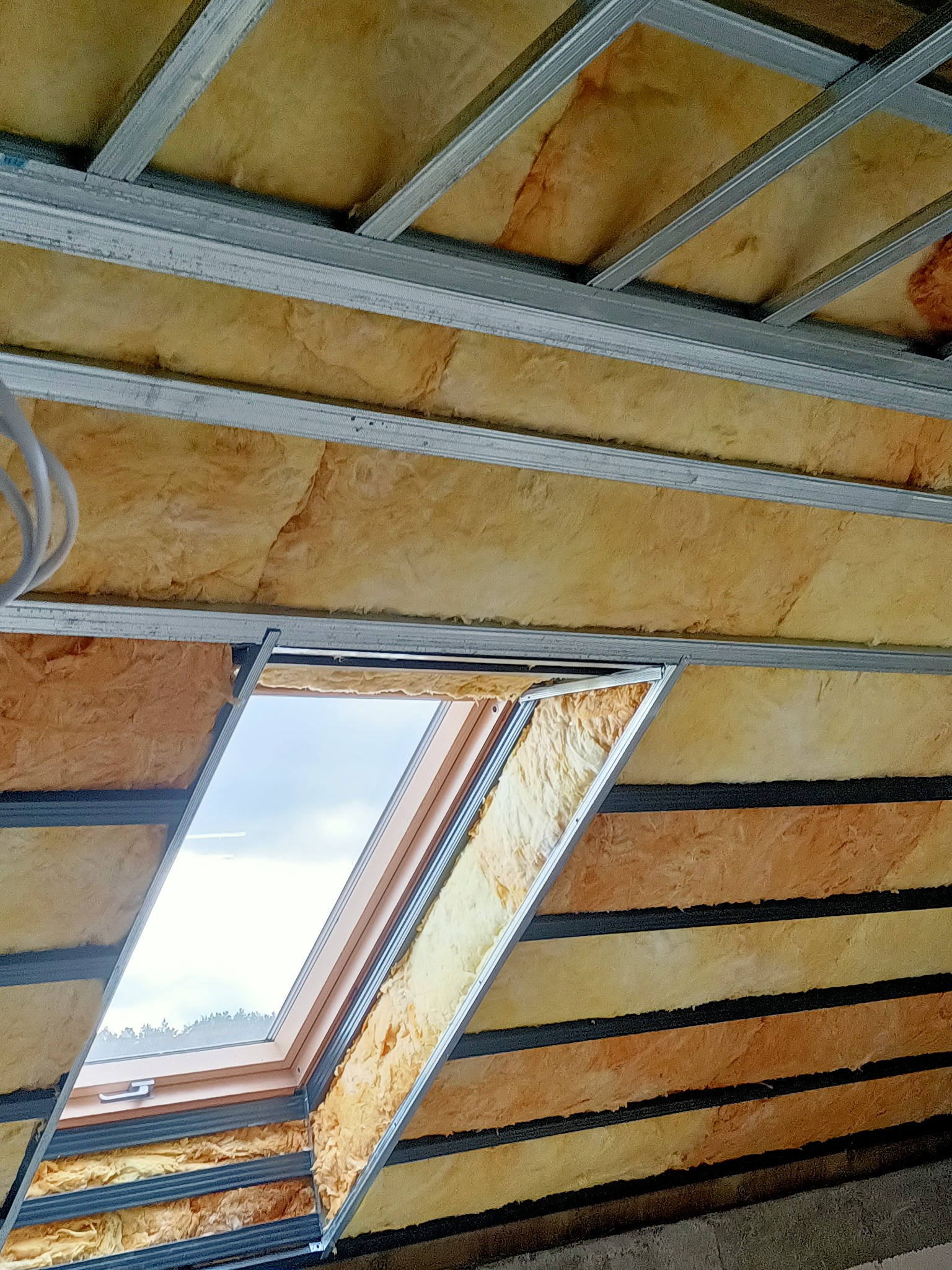 Montaż izolacji z wełny mineralnej poddasza z oknem dachowym, widoczne metalowe profile konstrukcyjne sufitu.