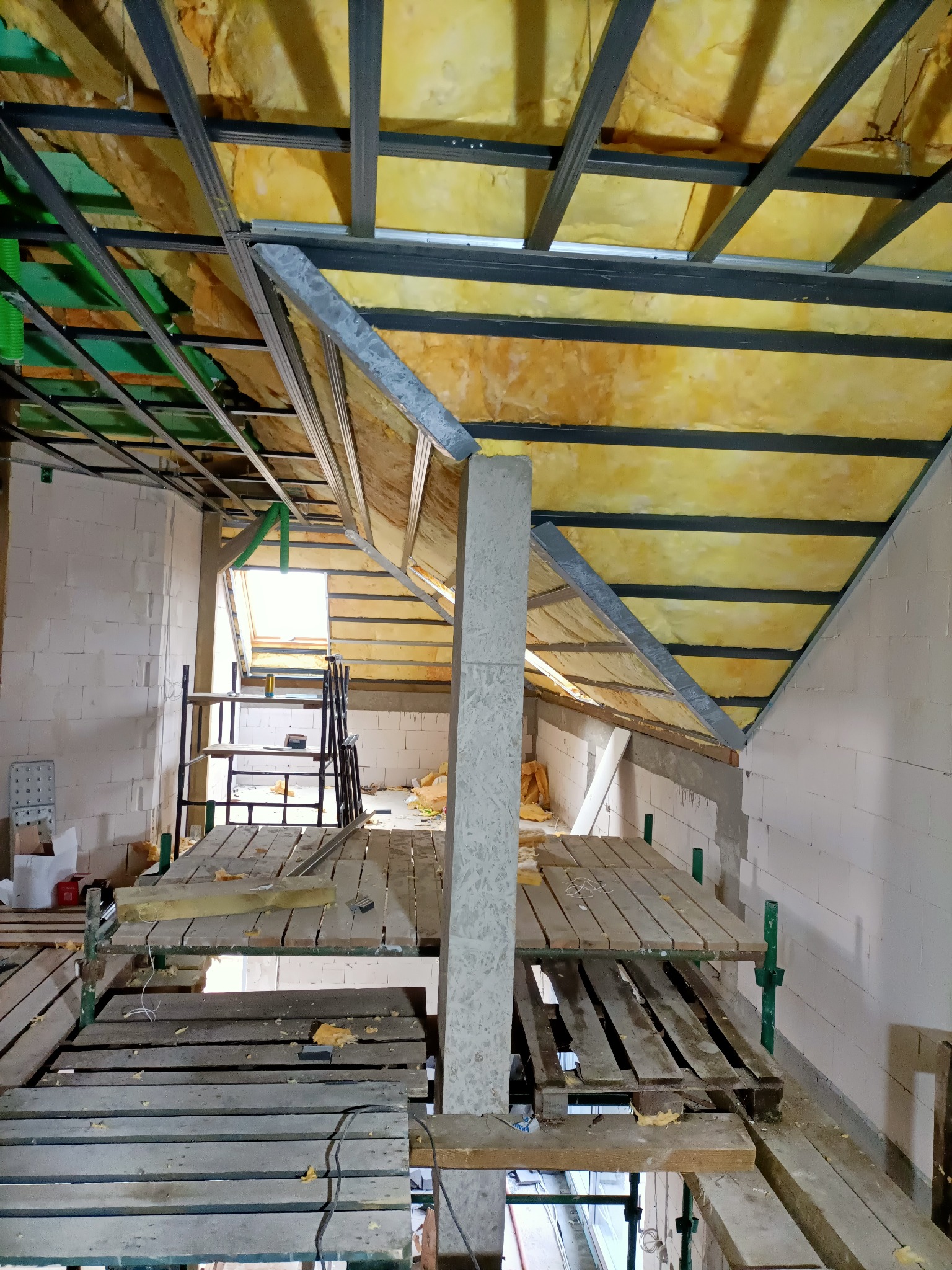 Montaż izolacji z wełny mineralnej na poddaszu, widoczne rusztowanie i konstrukcja metalowa pod płyty gipsowo-kartonowe.