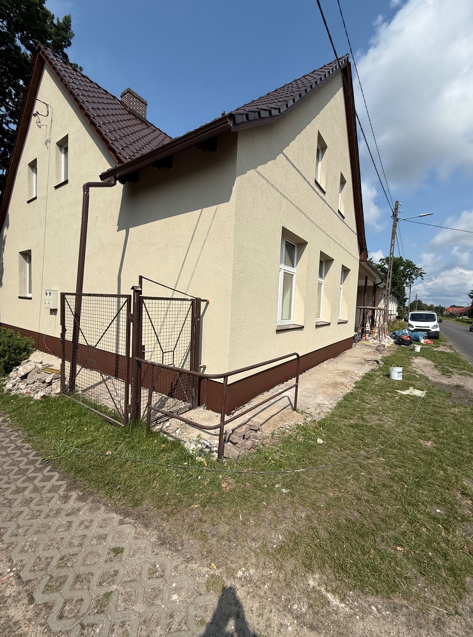 Jasny, otynkowany dom z brązowym dachem po renowacji. Widoczne prace wykończeniowe na zewnątrz, brama i fragment chodnika. Słoneczny dzień.