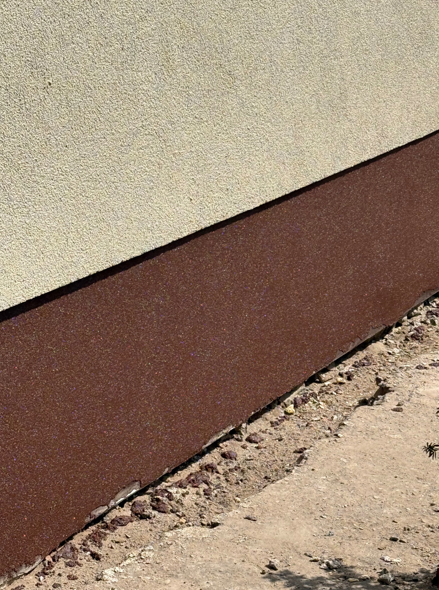Wykończona, brązowa cokolik elewacyjna z drobnym brokatem, przylegająca do beżowej, ziarnistej ściany. Widoczne połączenie z chodnikiem i fragment rośliny.