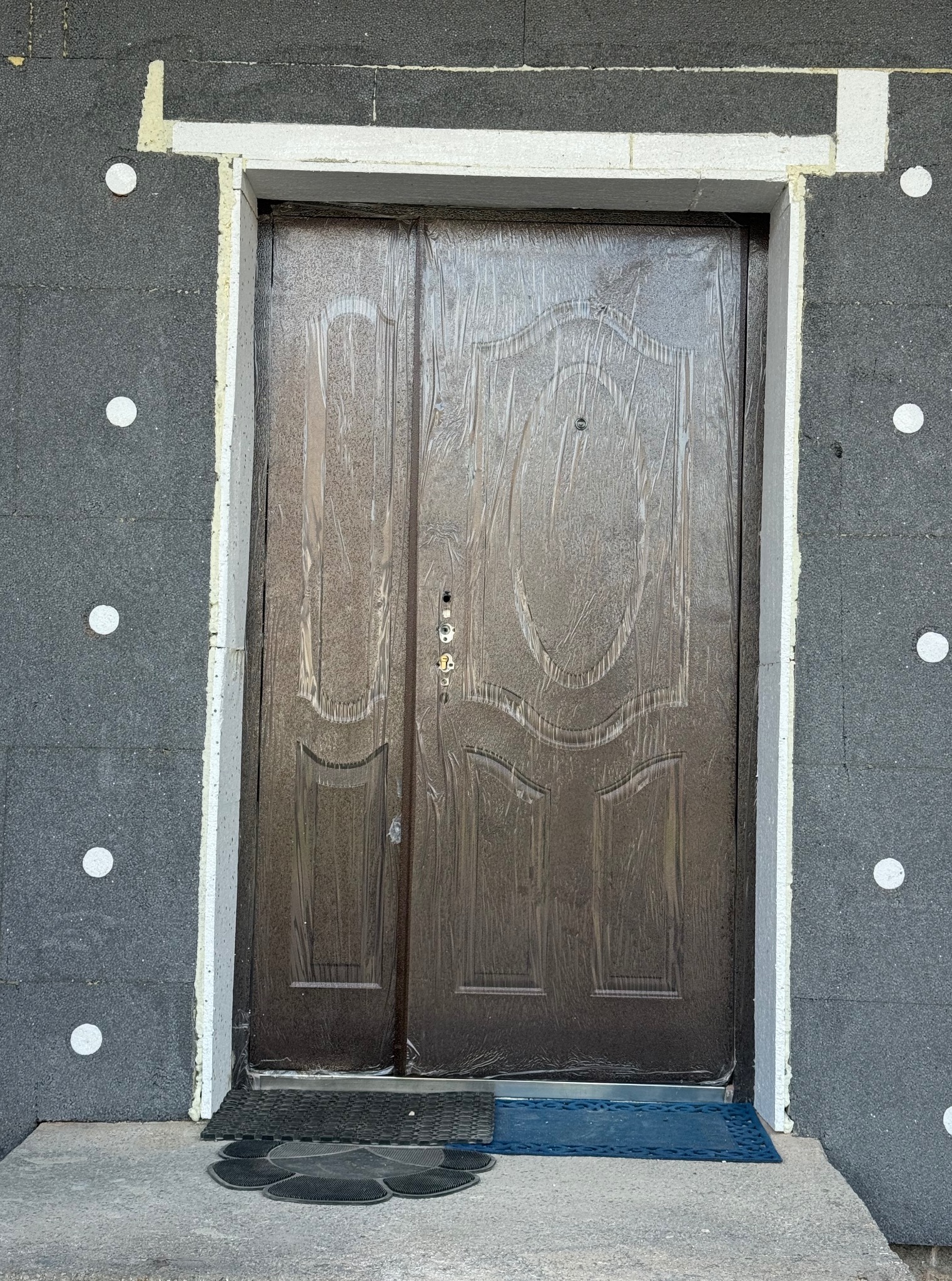 Ocieplona ściana budynku z brązowymi drzwiami wejściowymi, zabezpieczonymi folią ochronną, obok leżą wycieraczki. Widoczne prace wykończeniowe.