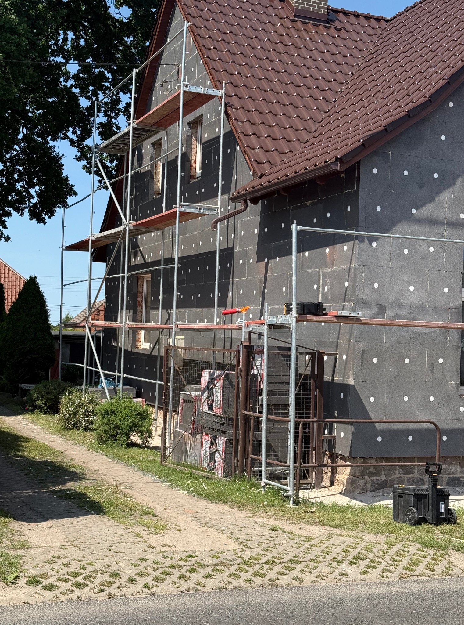 Dom w trakcie ocieplania. Ściany pokryte czarnym styropianem z białymi kołkami, rusztowania, brązowy dach. Widoczne materiały budowlane i skrzynka narzędziowa.