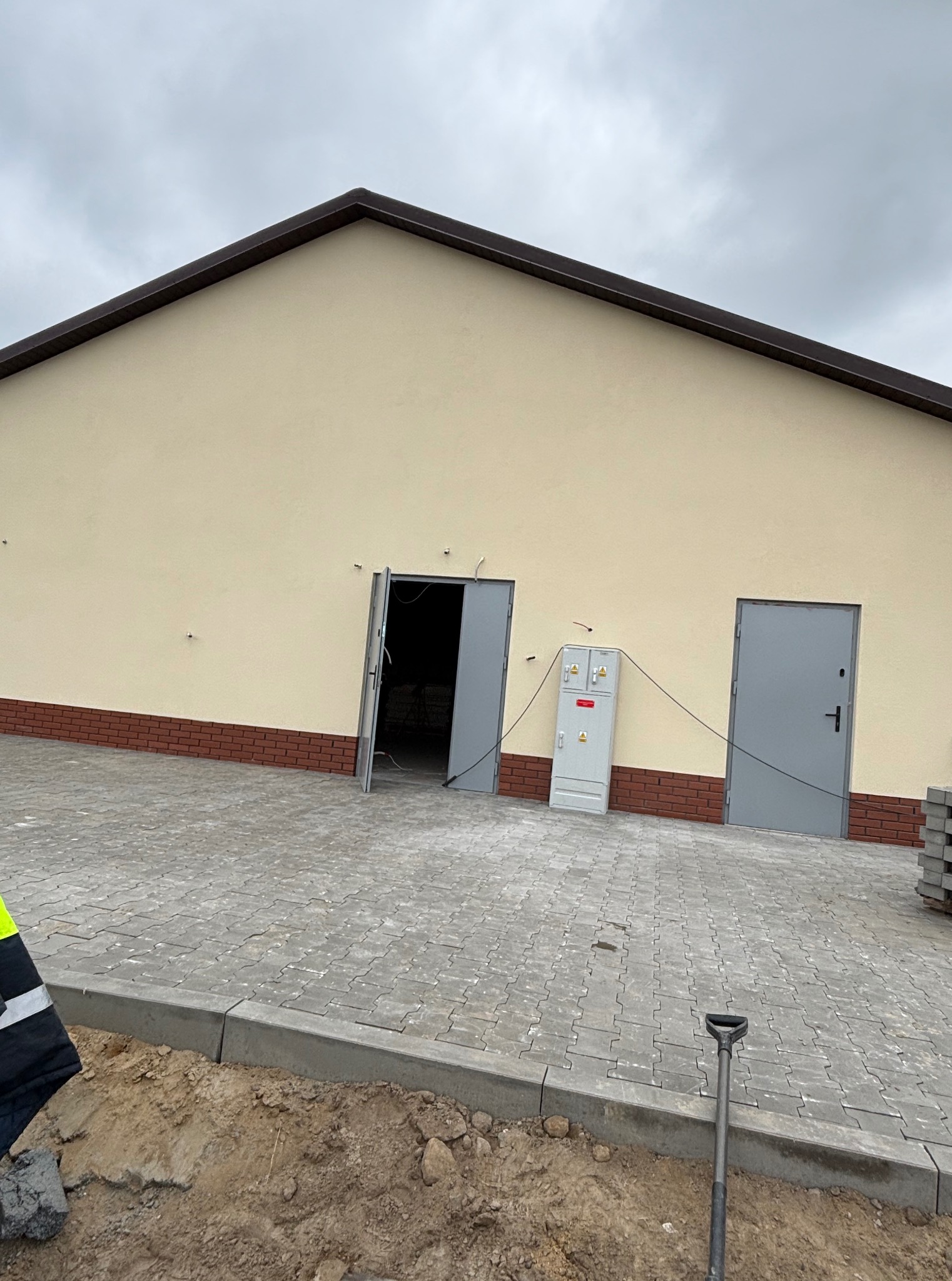 Elewacja budynku z beżową ścianą i ceglaną podmurówką. Widoczne dwoje szarych drzwi, skrzynka elektryczna, kostka brukowa i fragment łopaty w ziemi.