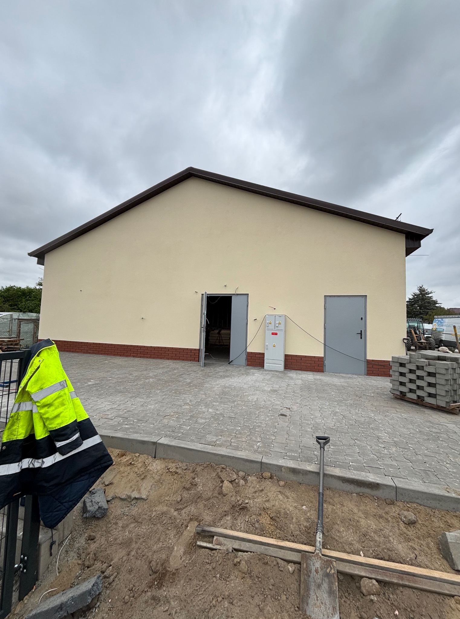 Budynek po malowaniu elewacji z widocznymi detalami: ceglana podmurówka, szara kostka brukowa, żółta kurtka i narzędzia sugerujące prace wykończeniowe na zewnątrz.