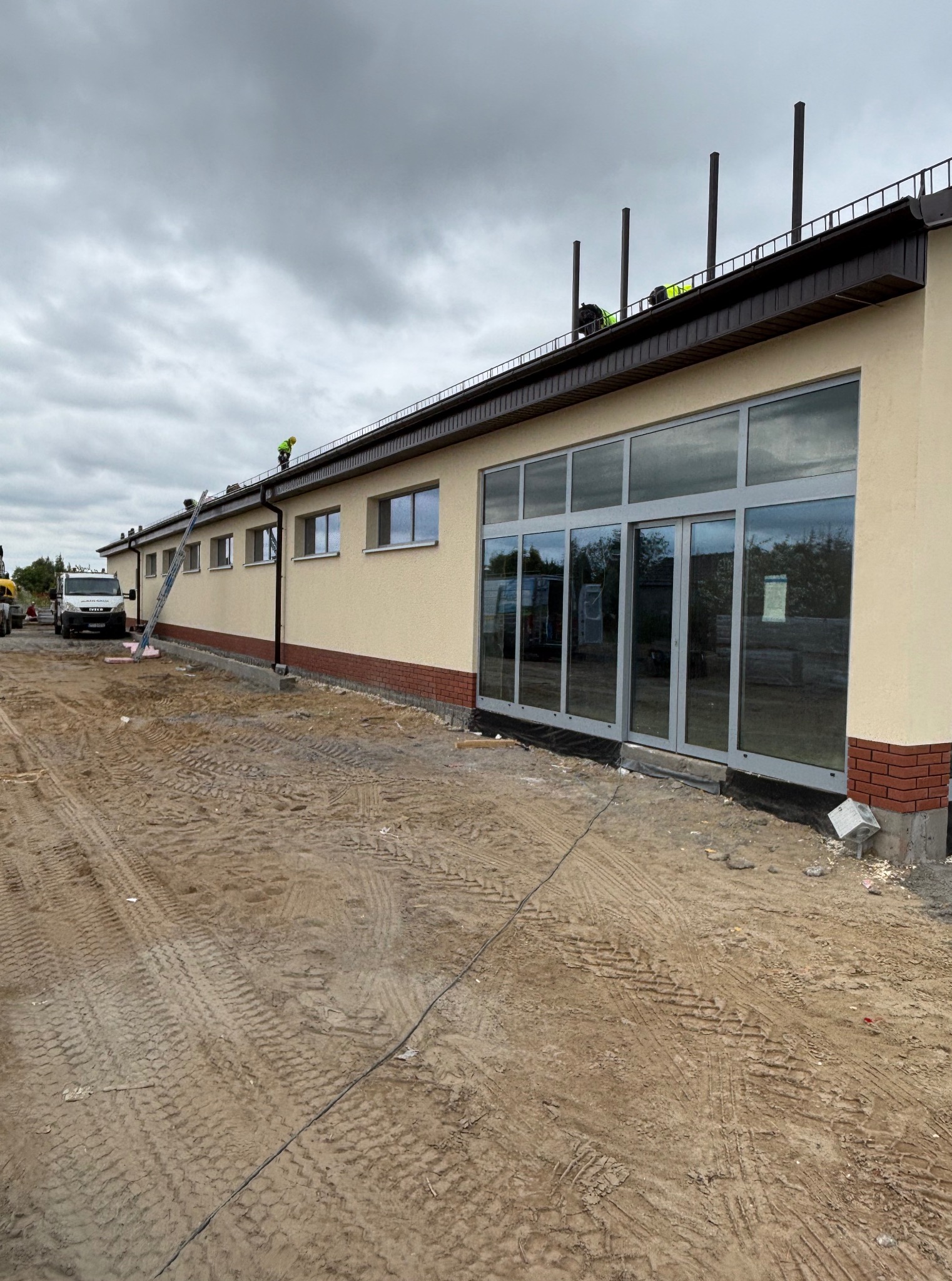 Budynek z beżową elewacją i dużymi oknami, z pracownikami na dachu, w trakcie prac wykończeniowych. Widoczny podjazd z piaskiem i ślady opon.