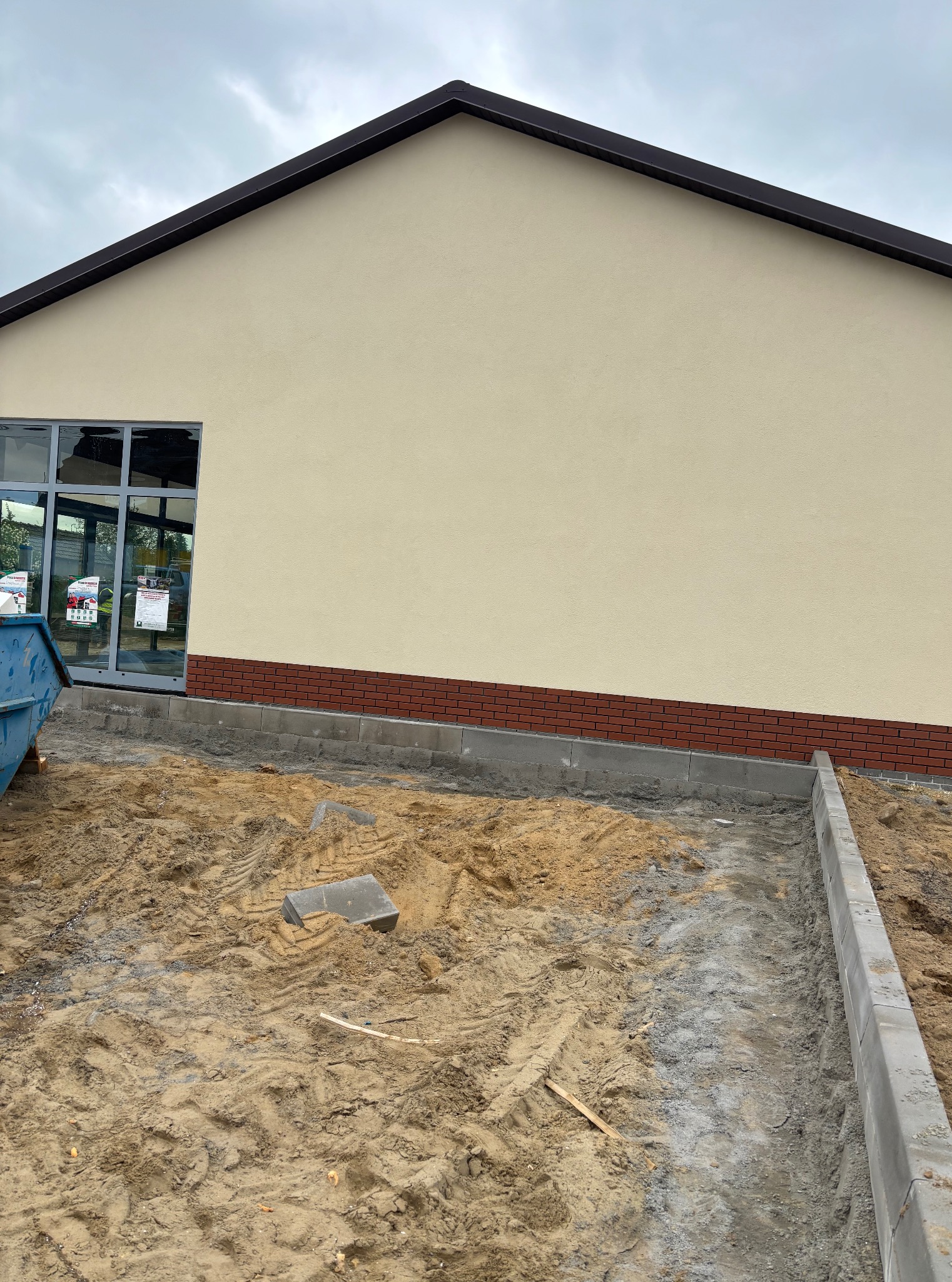 Elewacja budynku z oknem i ceglanym cokołem. Ułożone obrzeża chodnika z kostki betonowej i piasek. Widok na fragment budynku w pochmurny dzień.