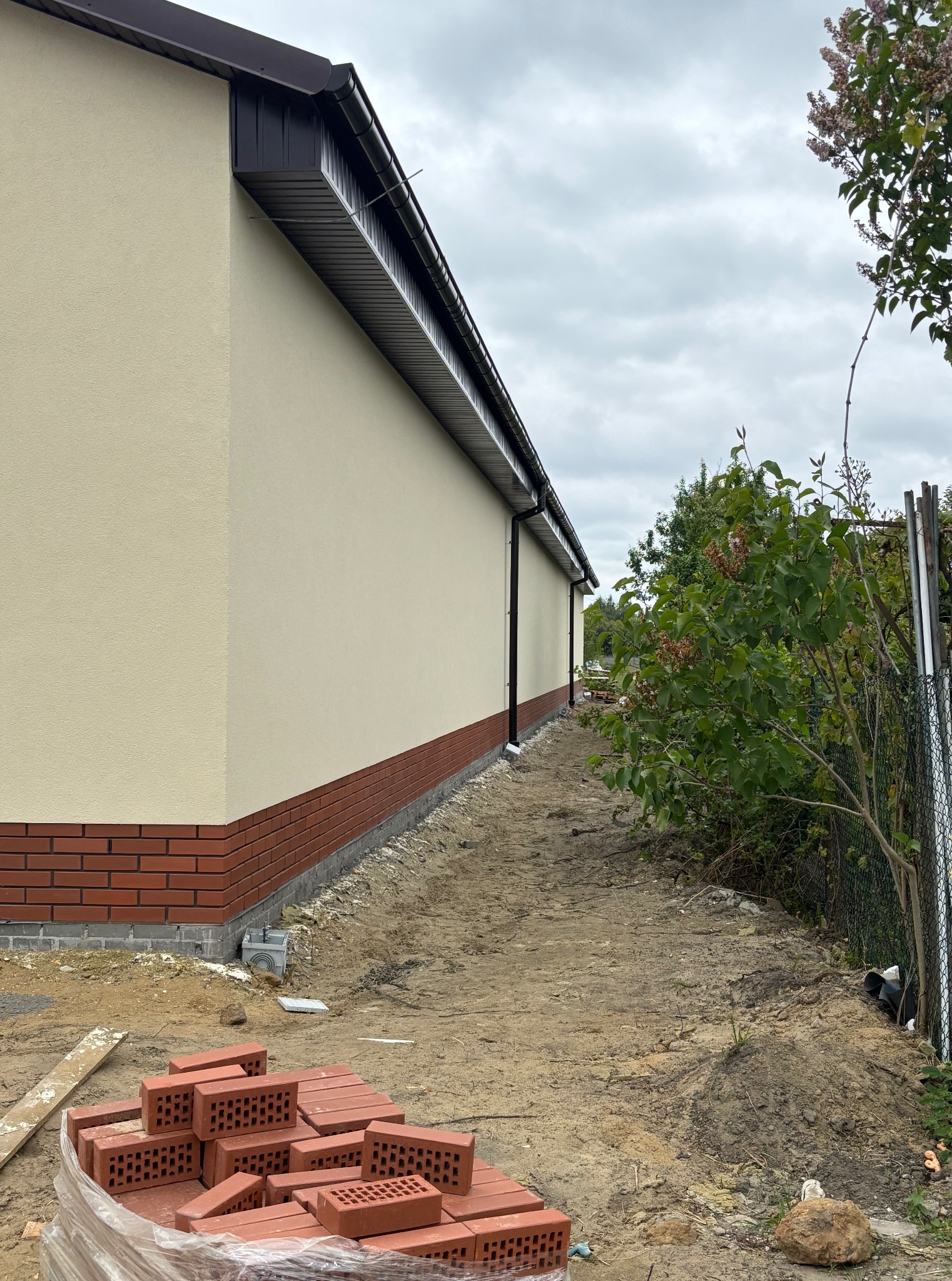 Elewacja budynku z beżowym tynkiem i czerwoną cegłą u podstawy. Widoczne rynny i stos cegieł. Teren wokół budynku przygotowany pod dalsze prace.