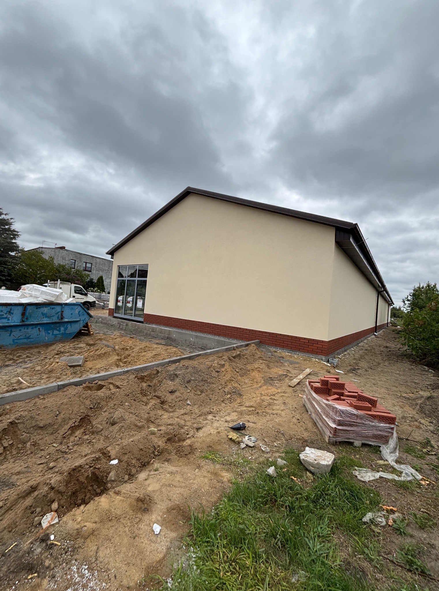 Otynkowany budynek z ceglaną podmurówką i szarą stolarką okienną w trakcie prac wykończeniowych na zewnątrz, widok z zewnątrz w pochmurny dzień.