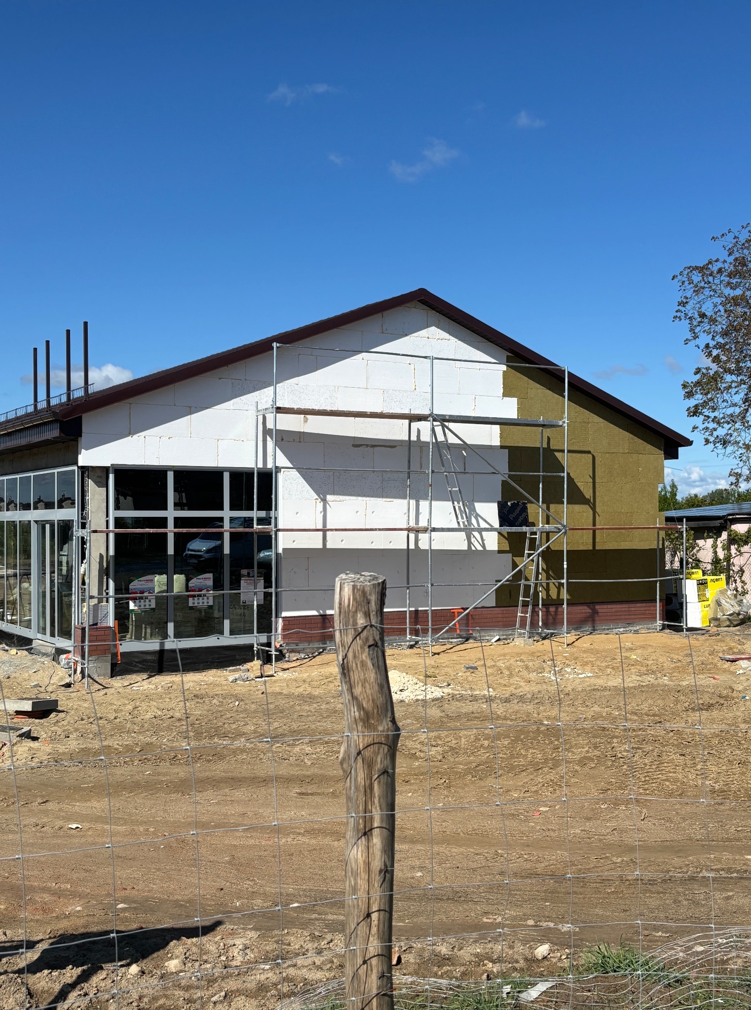 Ocieplanie budynku z rusztowaniem: część ściany z białym styropianem, część z brązową wełną mineralną. Widoczne okna i teren budowy.
