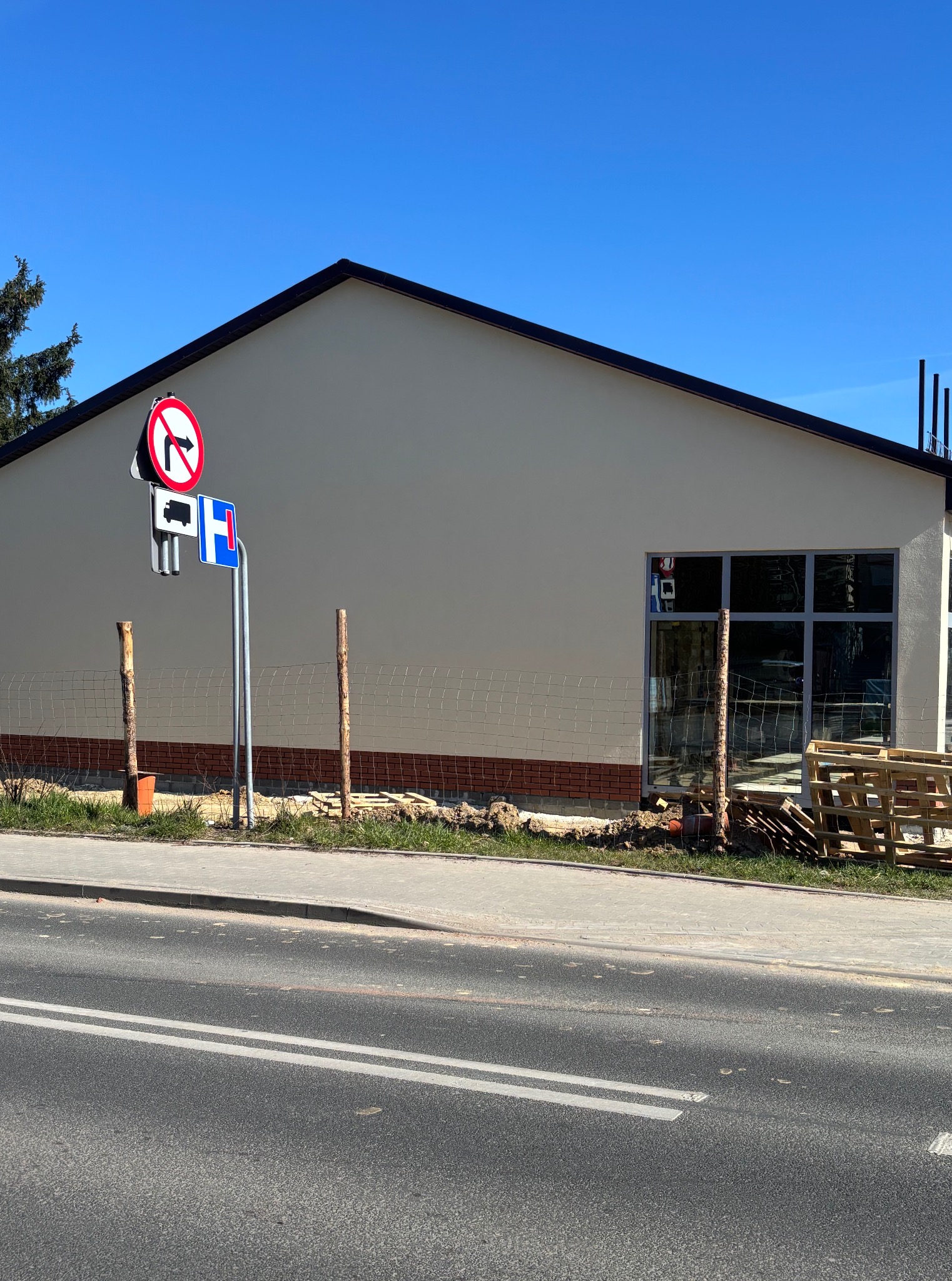 Budynek z beżową elewacją, częściowo wykończony czerwoną cegłą u dołu. Widoczne okna, znaki drogowe i palety. Prace wykończeniowe w toku.