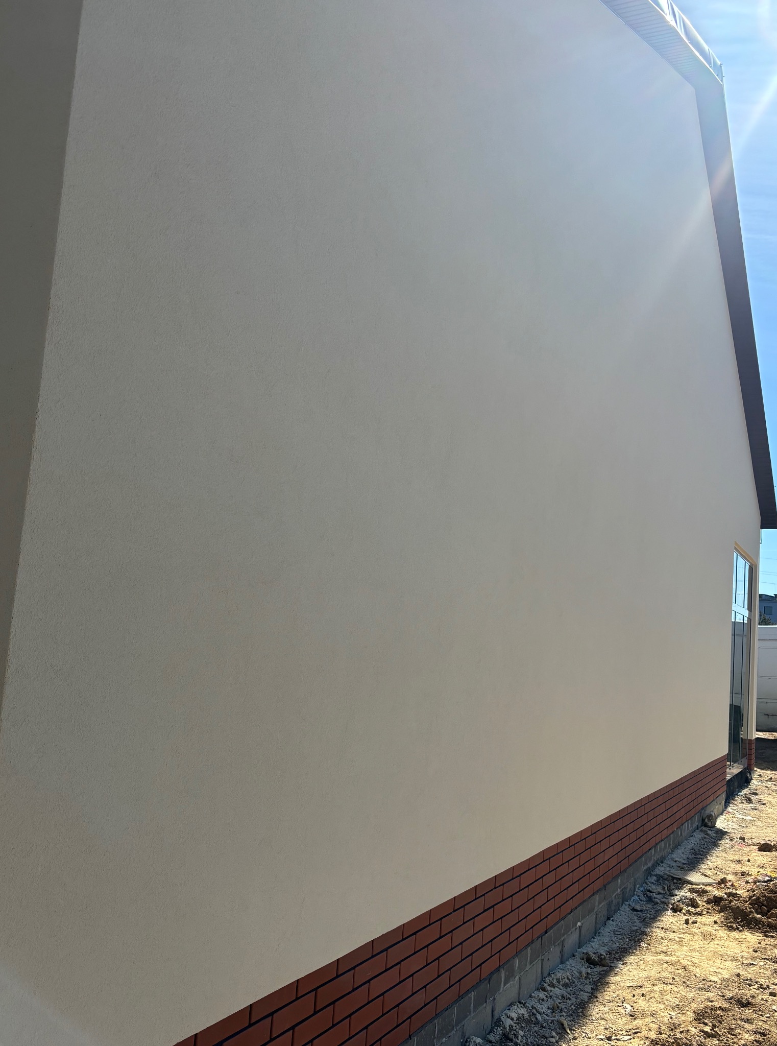 Ściana budynku po nałożeniu gładzi i pomalowaniu na beżowo, z widocznym ceglanym cokołem i fragmentem okna. Słoneczny dzień.