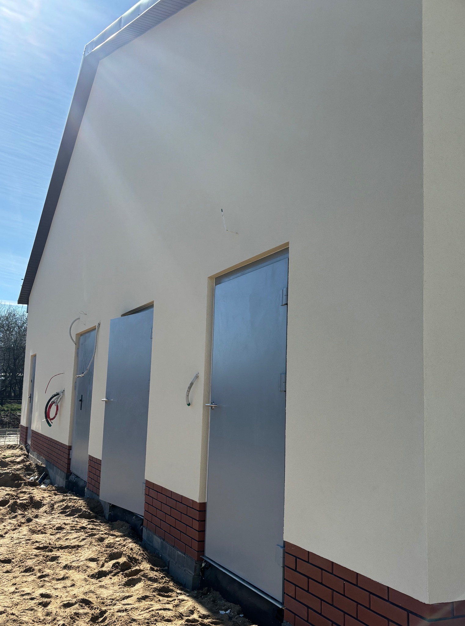 Otynkowana ściana budynku z trzema metalowymi drzwiami, wykończona czerwoną cegłą u podstawy i piaskowym podłożem. Widoczne przewody elektryczne.