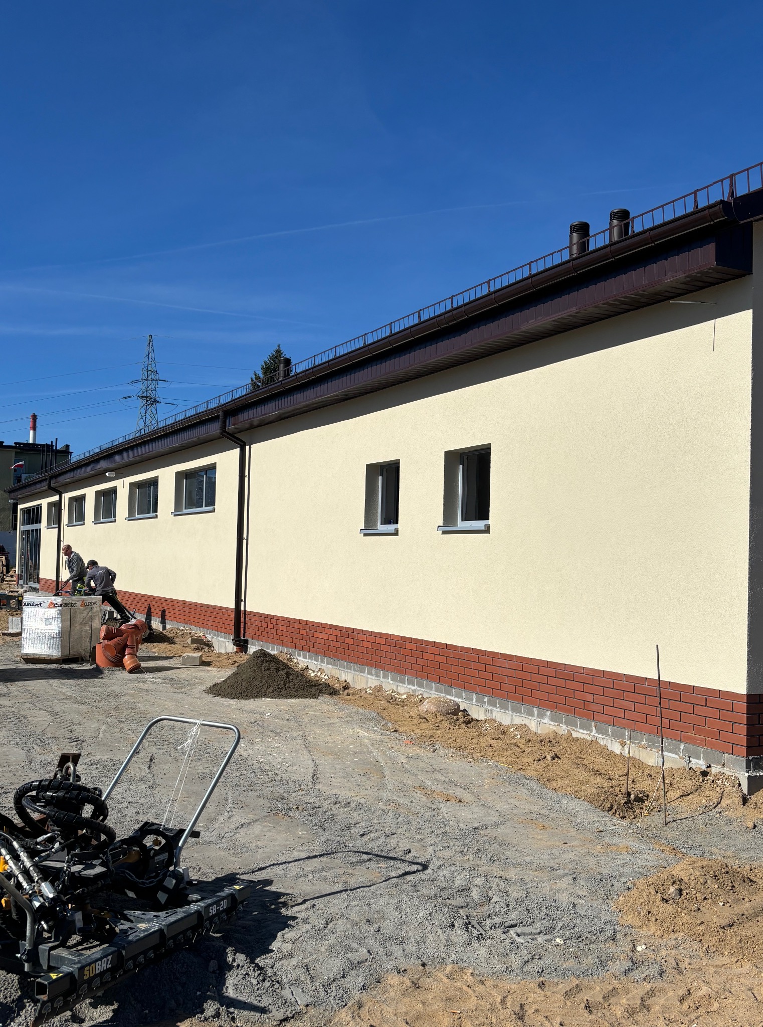 Prace wykończeniowe elewacji budynku: ocieplenie, ceglana podmurówka, żółta ściana. Na pierwszym planie ubijarka. Dwóch robotników przy pracy. Słoneczny dzień, błękitne niebo.