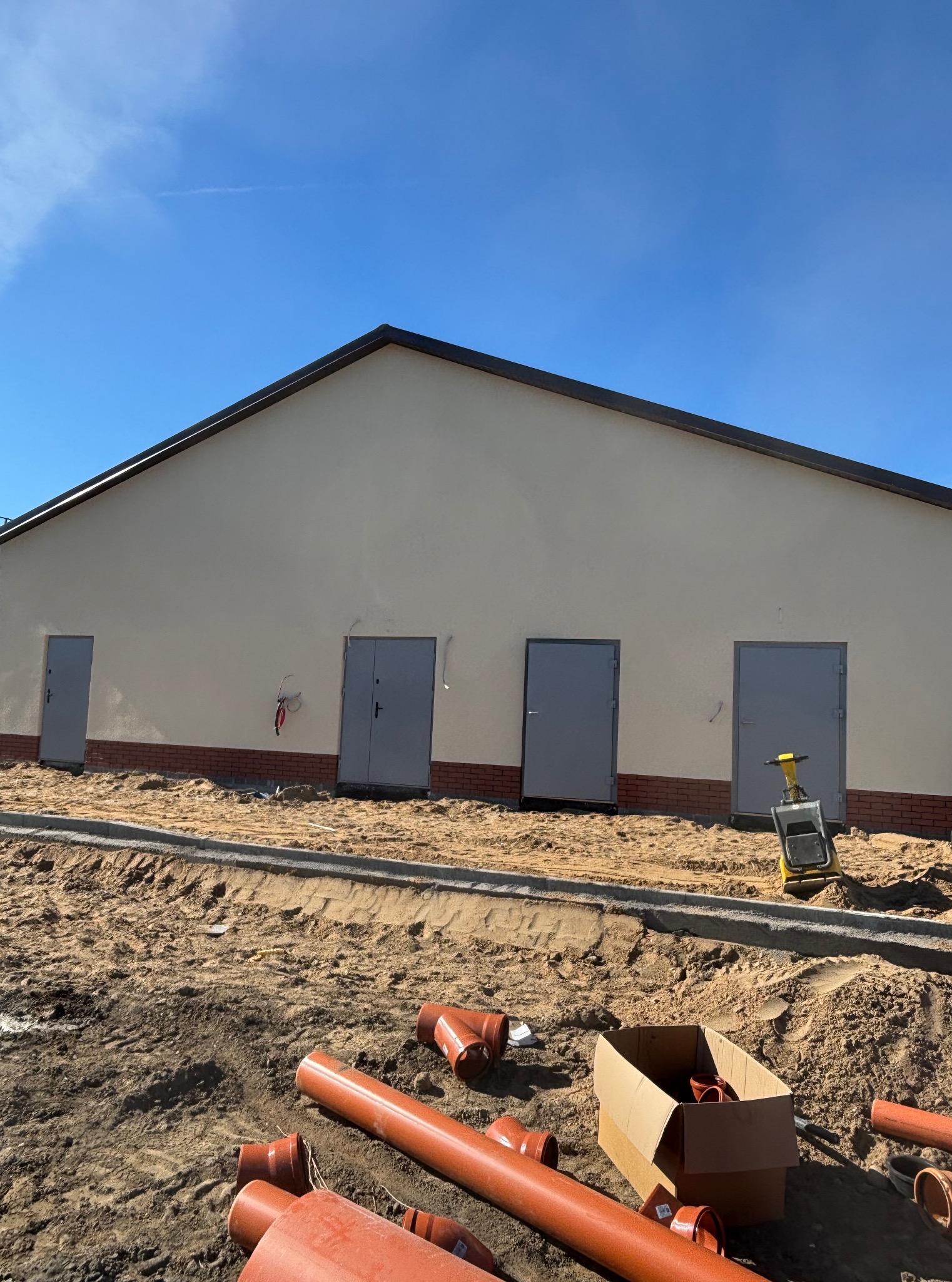 Budynek z czterema szarymi drzwiami i ceglaną podmurówką, widoczne pomarańczowe rury kanalizacyjne i ubijak wibracyjny. Prace ziemne w toku, błękitne niebo w tle.