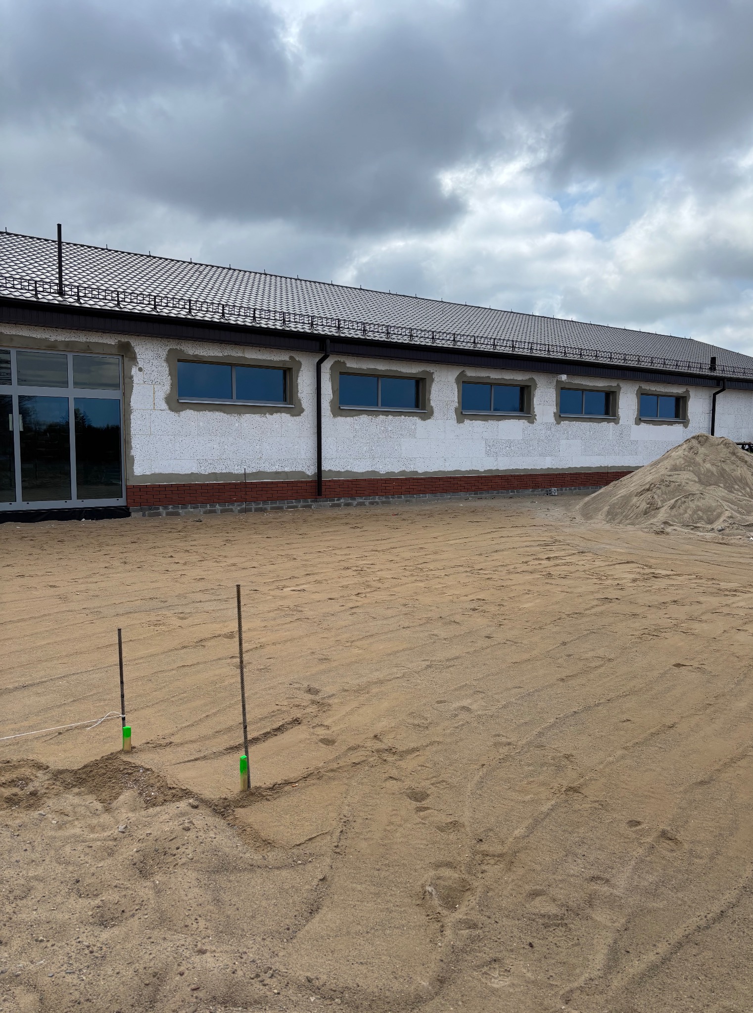 Budynek w trakcie wykończenia elewacji z widocznym piaskiem na zewnątrz, sugerujący prace ziemne lub przygotowanie terenu pod ogród.