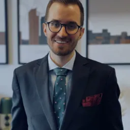 Uśmiechnięty prawnik w okularach i ciemnym garniturze, z bordową poszetką w kieszonce, na tle abstrakcyjnych obrazów w białych ramach.