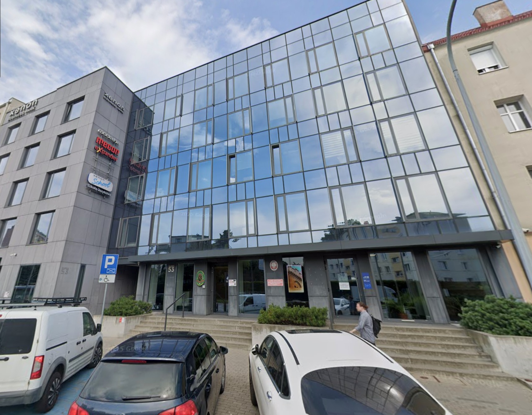 Nowoczesny budynek biurowy z przeszkloną fasadą, tabliczkami firmowymi i zaparkowanymi samochodami. Widoczny znak P dla niepełnosprawnych i numer budynku 53.