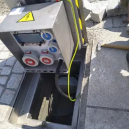 Otwarta skrzynka elektryczna z gniazdami przemysłowymi umieszczona w chodniku z kostki brukowej, widoczny kabel ochronny i znak ostrzegawczy o wysokim napięciu.