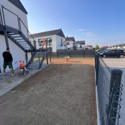 Maksymilian Gralec - Przygotowany teren pod trawnik w Kondratowicach. Widoczna gleba, obok stoi glebogryzarka, a w tle zabudowa szeregowa i zaparkowane samochody. Metalowe ogrodzenie.