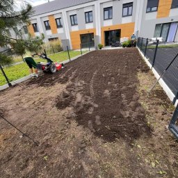 Maksymilian Gralec - Przygotowanie gruntu pod trawnik: gleba spulchniona glebogryzarką, widoczne ślady opon, w tle budynek mieszkalny z elementami drewnianymi.