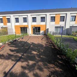 Maksymilian Gralec - Przygotowany teren pod trawnik z widocznymi liniami nawadniania, obok posadzone niskie krzewy, w tle budynek mieszkalny z drewnianymi elementami elewacji.