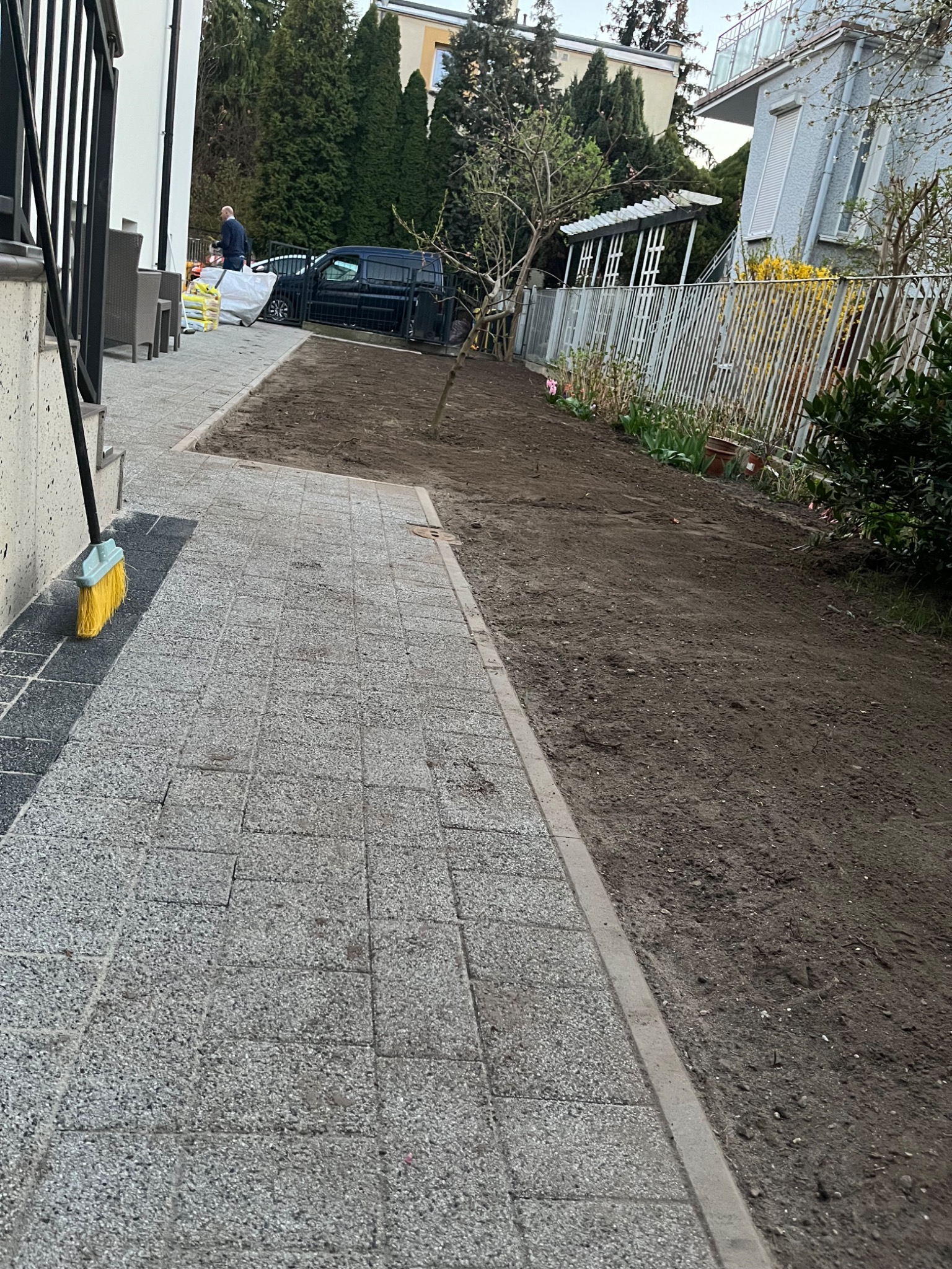 Przygotowany teren pod trawnik, widoczna świeżo przekopana ziemia, obok ułożona kostka brukowa, w tle zaparkowany samochód dostawczy i pracownik.